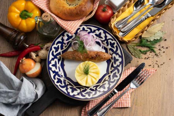 Un kebab grillé dressé sur une assiette orientale bleue et blanche, entouré de légumes frais et de pain traditionnel, typique d'une première destination gastronomique en Asie centrale.