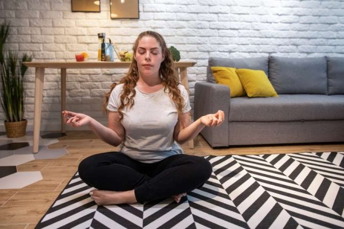 Femme assise en posture de méditation sur un tapis géométrique pour prendre soin de sa santé mentale dans un salon chaleureux et apaisé.