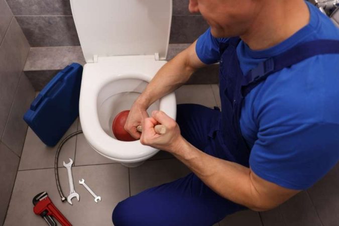 Un plombier en tenue bleue utilise une ventouse rouge pour déboucher un WC dans des toilettes carrelées.