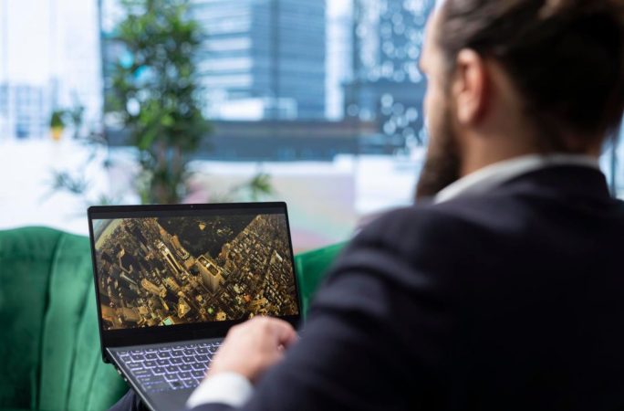 Un homme en costume analyse une vue aérienne urbaine sur son ordinateur portable pour consulter base dvf prix vente reel.